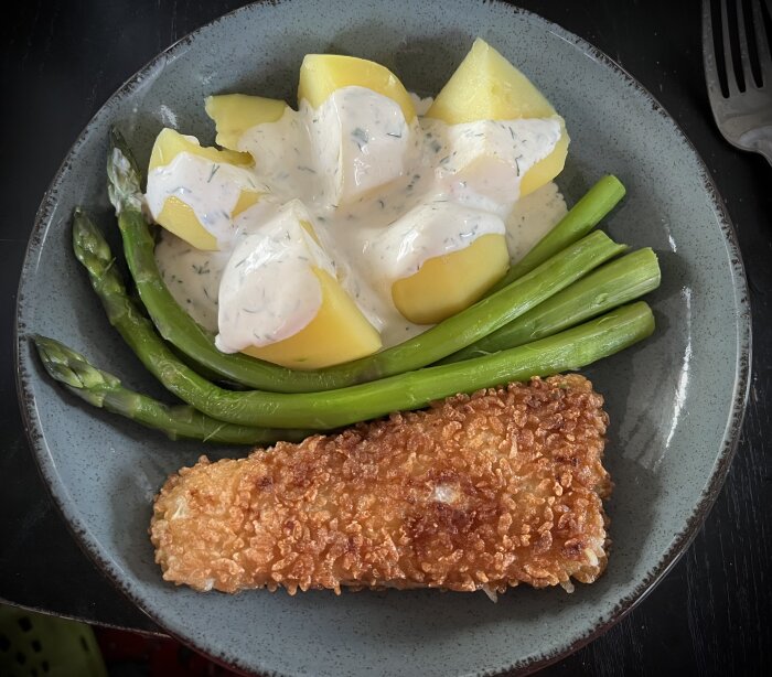 Panerad fisk med klyftpotatis och gröna sparrisar toppade med dill- och citronsås på en grå tallrik.