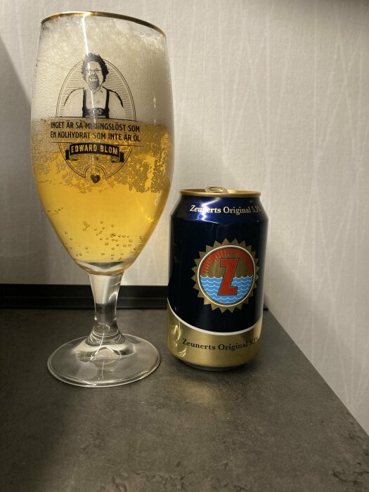 Ölglas halvfullt med skum och citat av Edward Blom, intill en oöppnad Zeunerts ölburk på bänk.
