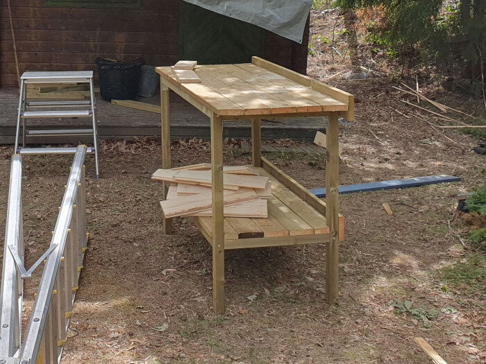 Hembyggt arbetsbord av trä med en tjockare skiva och robust underrede, bredvid ett mindre träbord, utomhus.