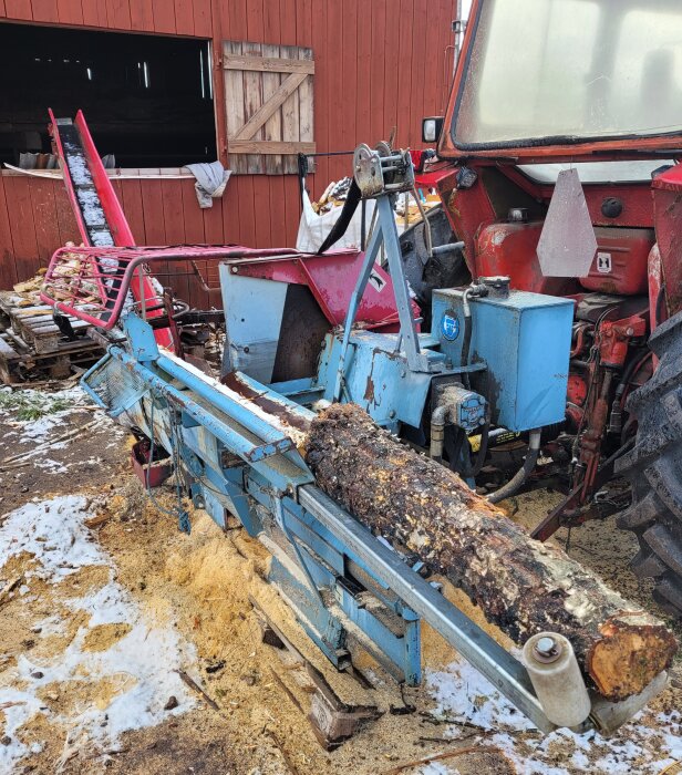 En logg placerad i en blå mobilsågbänk för sågning utomhus, med en röd traktor i bakgrunden.