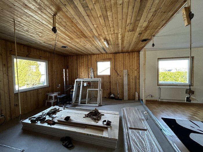 Rum under renovering med träpanel på väggar och tak, fönster, byggmaterial och verktyg utspridda.