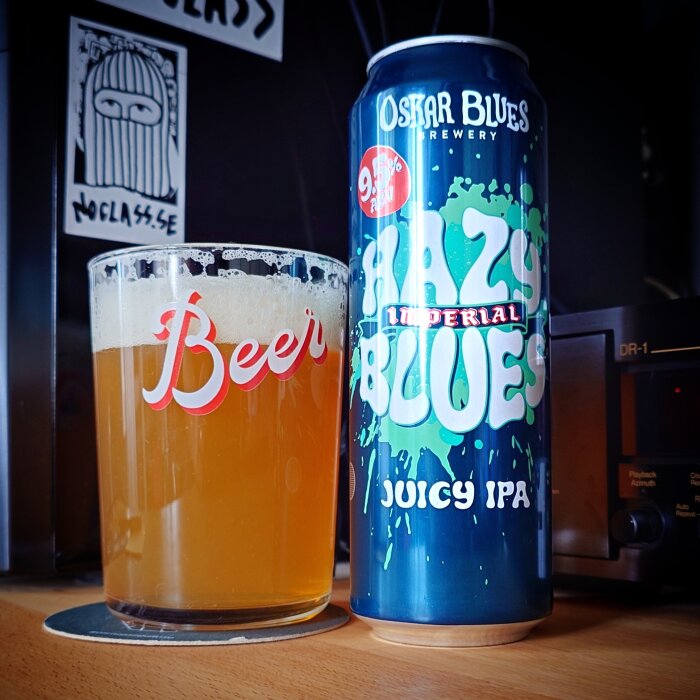 Ölglas med skum och en hög burk av Oskar Blues Juicy IPA på en bänk, med en beer-sticker i bakgrunden.