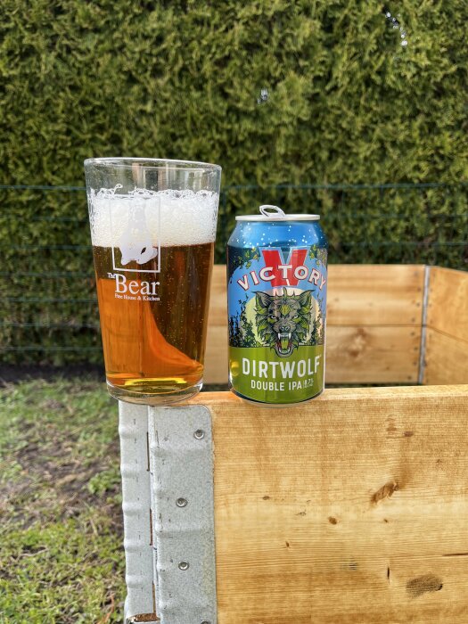 Ölglas på träkant med en burk Victory Dirtwolf Double IPA, omgiven av nya pallkragar och häck i bakgrunden.