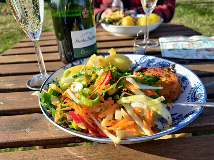 Middag utomhus med en tallrik med lax och grönsaker, ett glas champagne och en öppnad flaska Nicolas Feuillatte Organic.