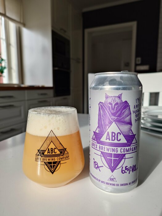 Ölglas med skum och Apex Brewing Company burk på ett bord i solljus.