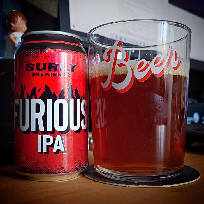 Burk Surly Furious IPA vid sidan av ett ölglas fyllt med bärnstensfärgad dryck, på ett skrivbord med dator.