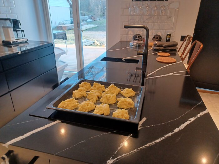 Plåt med oformade potatisbakelser på köksö i en modern köksmiljö, med utsikt till uteplats.