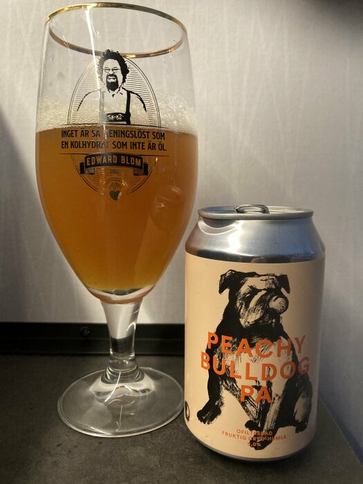 Ölglas med en ljus öl bredvid en burk med texten "Peachy Bulldog PA", inget skum och bakgrund.