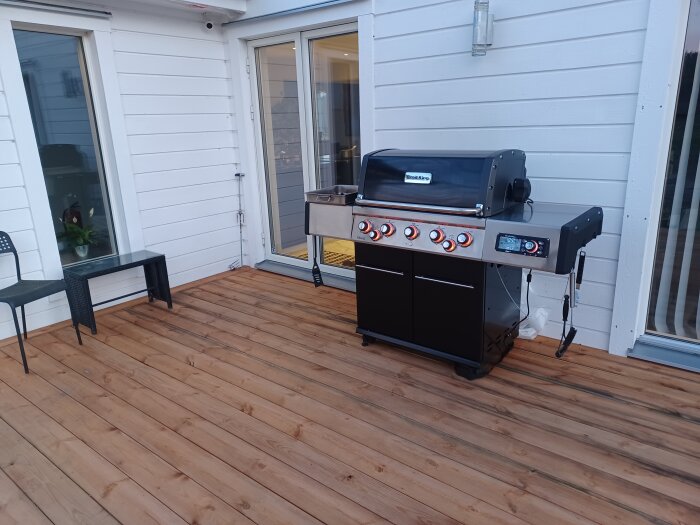 Modern gasolgrill på träaltan vid husvägg, redo för grillning.