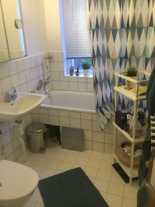 Äldre badrum med badkar, handfat, toalett och kakel, inrett med mönstrad duschdraperi och gröna accenter.