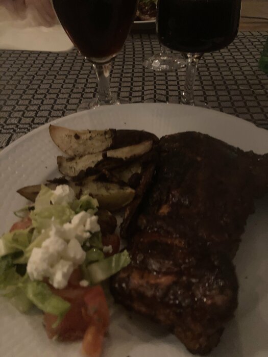 Grillad revbensspjäll på tallrik med klyftpotatis, sallad och två glas rödvin.