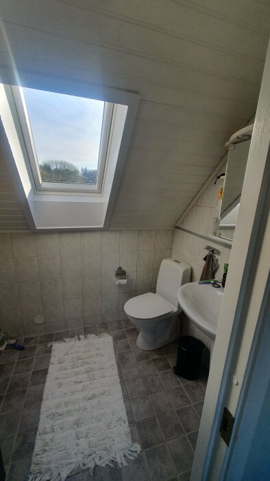 Badrum med snedtak, fönster i taket, toalett och handfat, potentiell plats för badrumsfläkt.