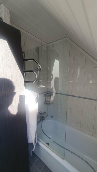 Badrum med badkar och duschskärm, låg vägghöjd, diagonalt infallande ljus, skugga av en person.