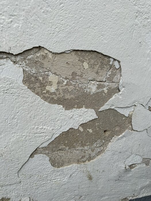 Sliten putsfasad med flagande vit färg som avslöjar underliggande brungrått lager på en äldre byggnad.