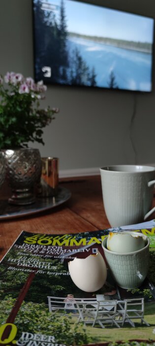 Ett köksbord med tidskrift, uppbrutet ägg i äggkopp, kaffekopp och suddig TV i bakgrunden.