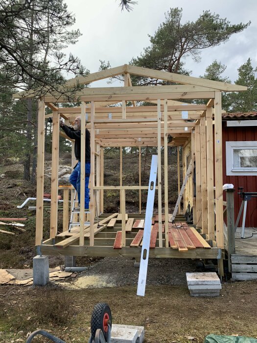 Person bygger trästomme till hus i skogsmiljö med verktyg och material runtomkring.