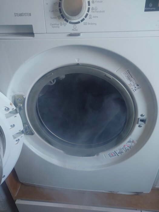 Tvättmaskin med öppen lucka visar rök och potentiellt skev trumma, ingen synlig skada eller felkod.