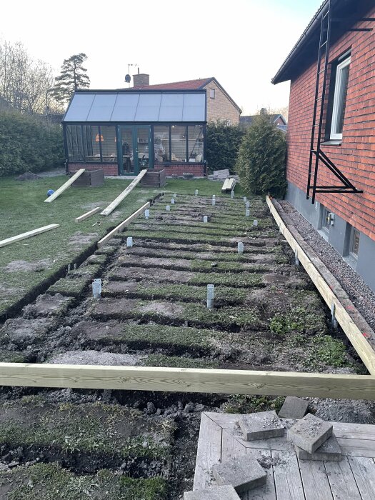 Grävt område för altanbygge med placerade markskruvar och en upplagd träregel i en trädgård bakom ett hus.