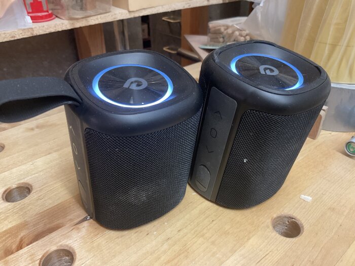 Två blåtandshögtalare på ett arbetsbord i en verkstad, lysande ringar runt knapparna indikerar att de är på.