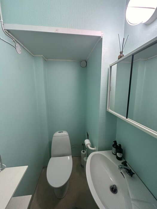 Toalett med stor avstånd från väggen, handfat, spegel och synliga rör i ett badrum behövande renovering.