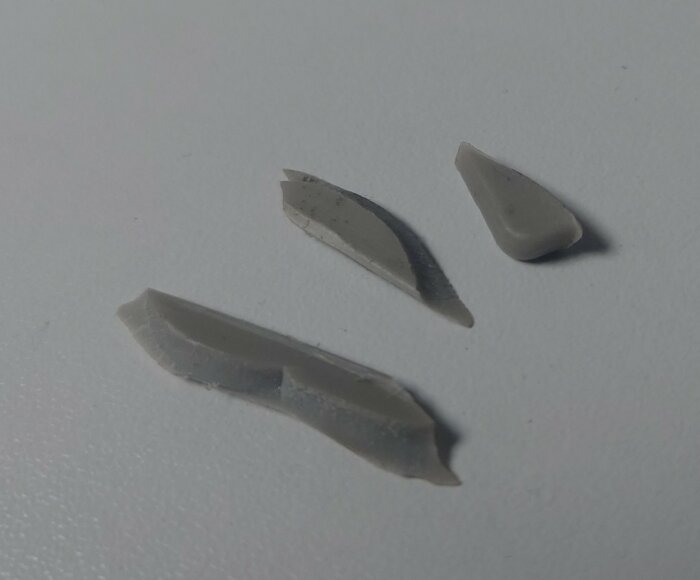 Tre grå plastbitar av olika former och storlekar på en vit bakgrund, misstänkta delar från en tvättmaskin.
