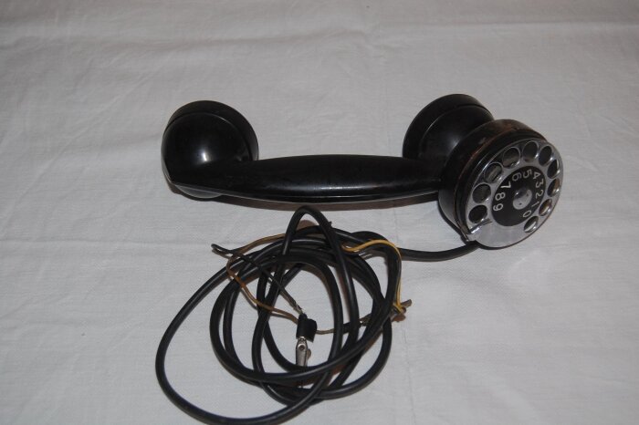 Svart, gammaldags telefon med vridskiva och sladd på vit bakgrund.