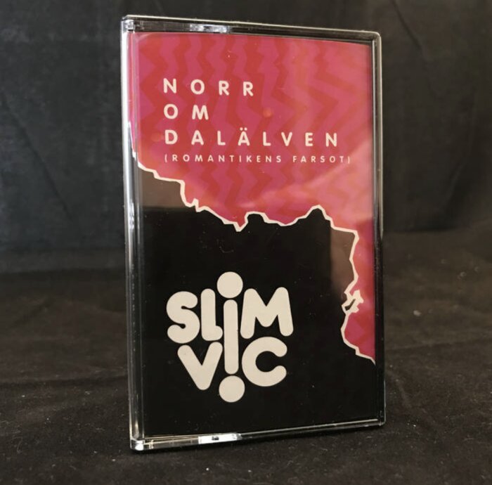 Kassettband med texten "NORR OM DALÄLVEN" och "SLIM VIC", svart bakgrund och rosa vågmönster.
