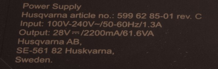 Etikett på en Husqvarna strömförsörjning med artikelnummer och specifikationer för ströminput och -output.