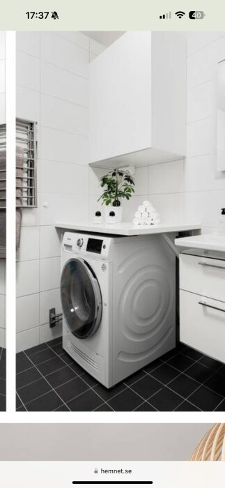 Kombi-tvättmaskin i badrum med vit arbetsbänk ovanför, vita väggar och mörkt klinkergolv.