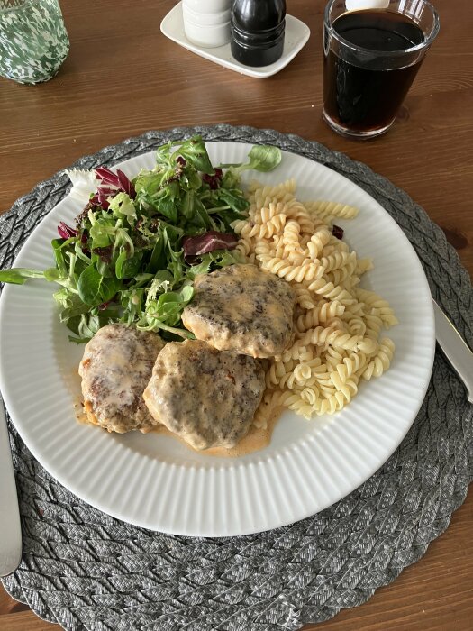 En måltid med fetaost- och tomatbiffar i en gräddig sås serverade med pastaskruvar och en grönsallad, samt ett glas mörk dryck.