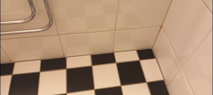 Hörnet av ett badrum med ny silikonfog mellan vita väggkakel och svartvita golvklinker.
