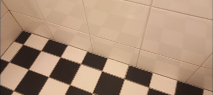Svartvitt rutigt badrumsgolv som möter en vägg med vita kakelplattor där en ny silikonfog har applicerats i vinkeln.