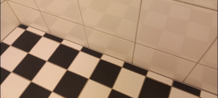 Hörn av ett badrum med ny silikonfog mellan vita väggkakel och svartvita golvklinker.
