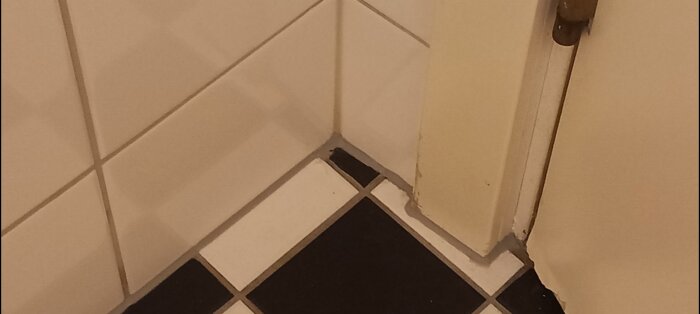 Hörnet av ett kaklat badrum med ny silikonfog mellan svartvitt golv och vita väggar, med vissa ojämnheter synliga.