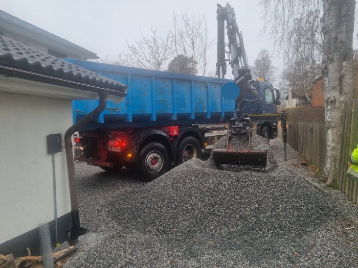 Lastbil med grävmaskinsarm som flyttar grushög bredvid en byggnad, arbetare syns delvis.