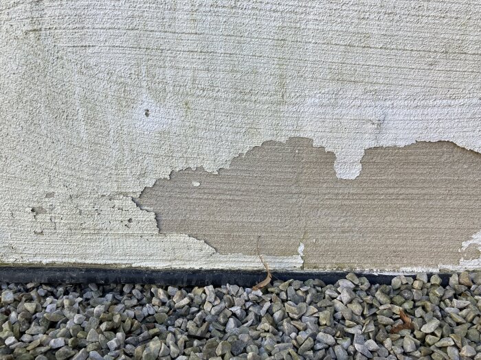 En närbild på en vägg med flagande färg ovanför en grusplan, eventuell KC- eller cementputs synlig.