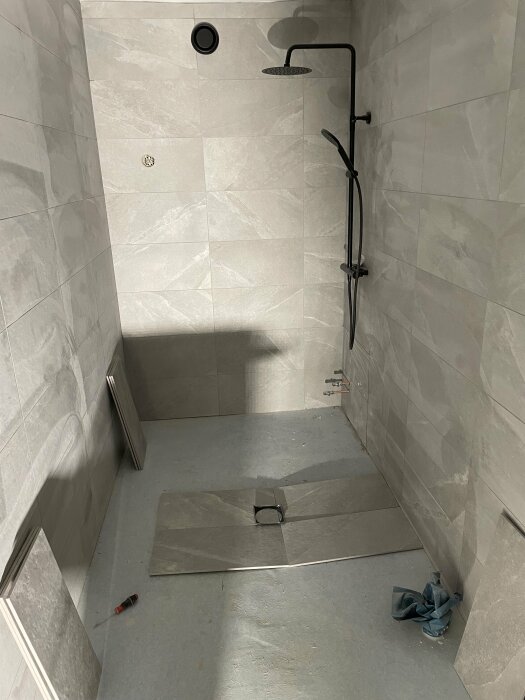 Ofärdigt badrum med gråa klinkerplattor på väggarna och på golvet framför en duschkabin, vissa ej installerade.