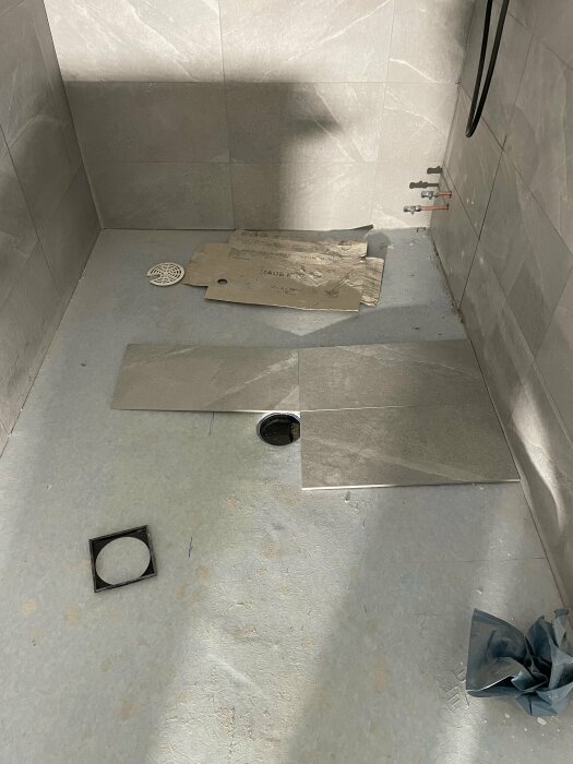 Ofärdigt badrumsgolv med påbörjad läggning av gråa 60x30 klinkerplattor, material och verktyg synliga.