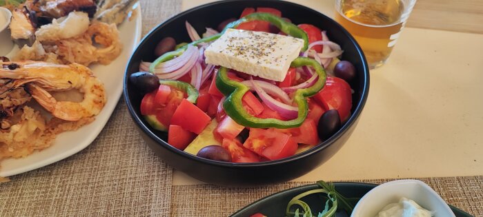 Grekisk sallad med fetaost, oliver och grönsaker, bakgrund med skaldjur och ölglas.