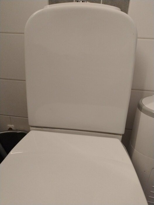 Stängd vit toalettstol med synligt läckageproblem vid ryggstödet i ett kaklat badrum.
