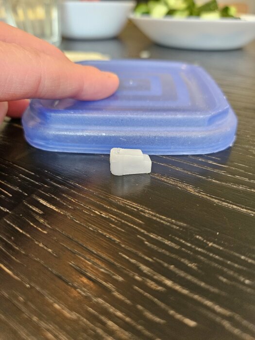 Hand håller ett litet vitt spröjsclip på ett mörkt bord med synlig textur.