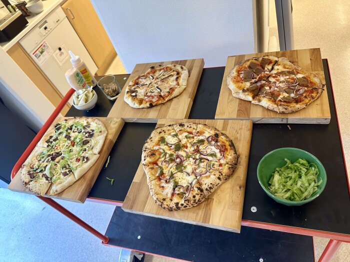 Fyra olika hemlagade pizzor på träbrädor med en skål sallad på ett köksbord.