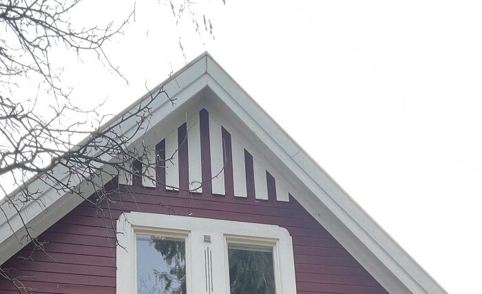 Gavel med randig ventilerad vind på röd träfasad och vita fönsterkarmar under bar gren.