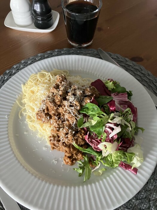 Spaghetti med köttfärssås och riven ost, sallad på sidan, salt- och pepparkar och ett glas mörk dryck i bakgrunden.