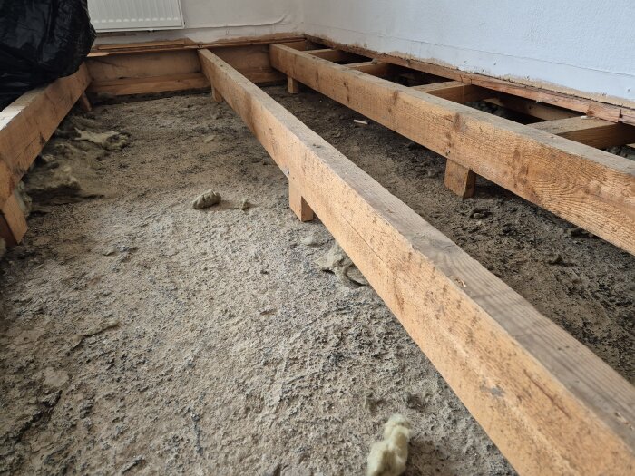 Delvis upprivet golv i enplanshus med synliga reglar av trä ovanpå en betongplatta och gammal isolering utspridd.