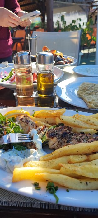 Souvlaki med pommes frites och tzatziki på en tallrik, blåmusslor i bakgrunden, middagsmiljö utomhus.