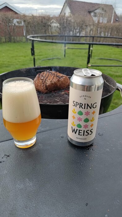 Ett glas öl och en ölburk märkt "SPRING WEISS" med grillat kött i bakgrunden och en trädgård.