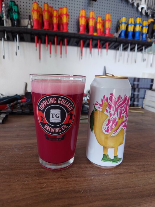 Glass med rosa dryck från Toppling Goliath Brewing och burk med tecknad design på bänk, verktyg i bakgrunden.