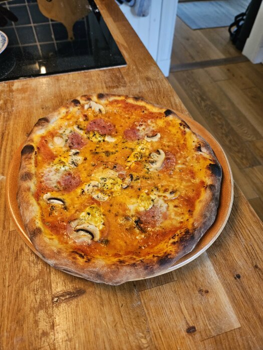 Hembakt pizza på inbränd pizzasten med salami, svamp och gul ost på ett träbord.