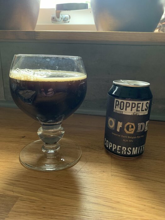 Ett glas mörkt öl bredvid en öppnad burk från Poppels bryggeri på ett träbord.